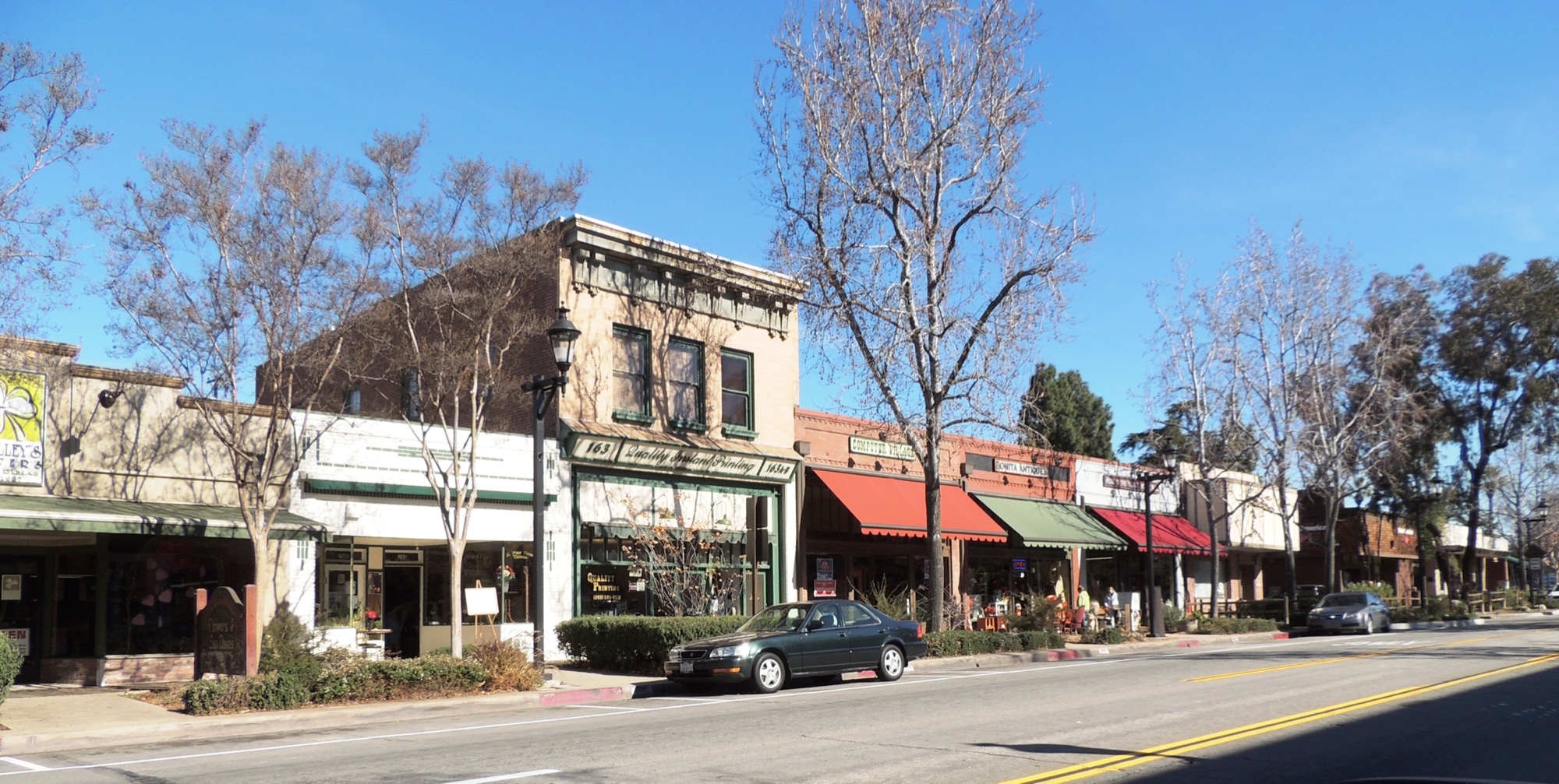 A daytime scene of a city street with colorful stores and parked cars.