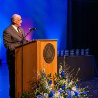 A man in a suit speaks at a podium decorated with a floral arrangement of blue and white flowers during an awards ceremony. Behind him, a table displays multiple glass awards against a backdrop lit with blue and purple lights.