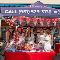 A group of six smiling women stand behind a booth at the San Dimas Beer and Wine Walk 2025. The booth, decorated with red bandana bunting and a blue canopy displaying a phone number and realtor’s name, showcases raffle baskets wrapped in cellophane. The sunny outdoor scene captures a friendly, community vibe.