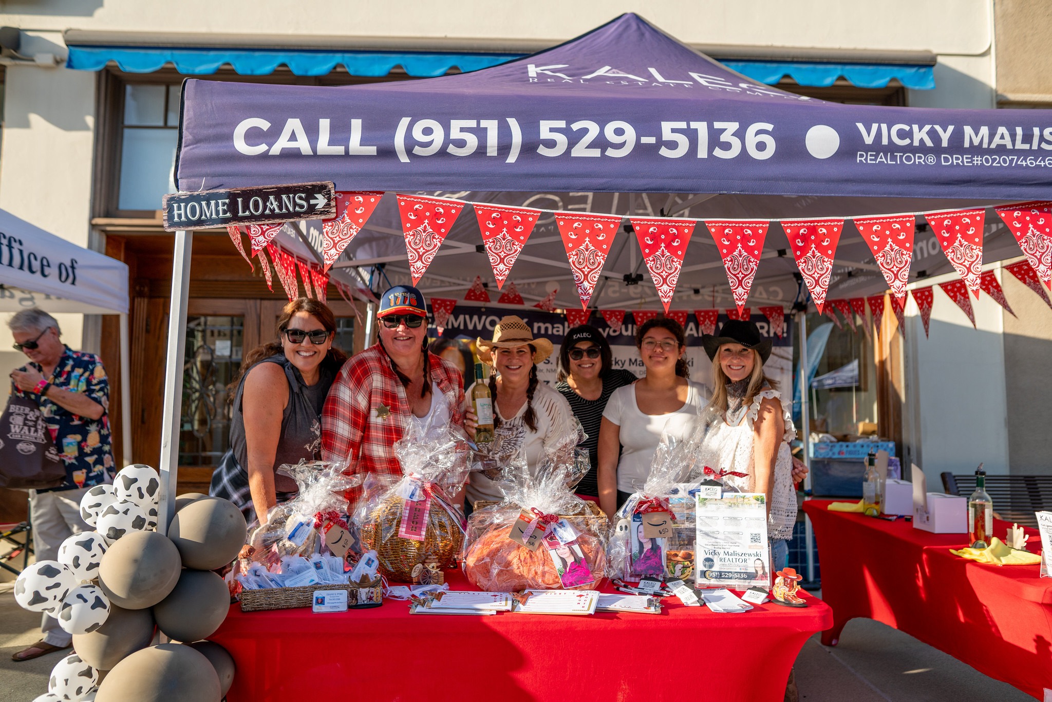 A group of six smiling women stand behind a booth at the San Dimas Beer and Wine Walk 2025. The booth, decorated with red bandana bunting and a blue canopy displaying a phone number and realtor’s name, showcases raffle baskets wrapped in cellophane. The sunny outdoor scene captures a friendly, community vibe.